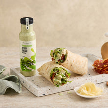 Load image into Gallery viewer, A bottle of Salad Servers Herb &amp; Caesar Dressing stands on a stone serving board next to two cut wrap burritos filled with greens and bacon, with a small dish of cheese and a cloth on a beige tabletop.
