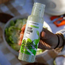 Load image into Gallery viewer, A hand holding a bottle of Salad Servers freshly dressed herb caesar dressing, with a bowl of greens blurred in the background.
