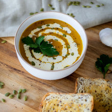 Load image into Gallery viewer, Bowl of pumpkin soup with cream swirl, seeds, and parsley, served with bread on a wooden board.
