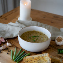 Load image into Gallery viewer, Bowl of creamy potato and leek soup garnished with herbs, served with bread, garlic, and candlelight.
